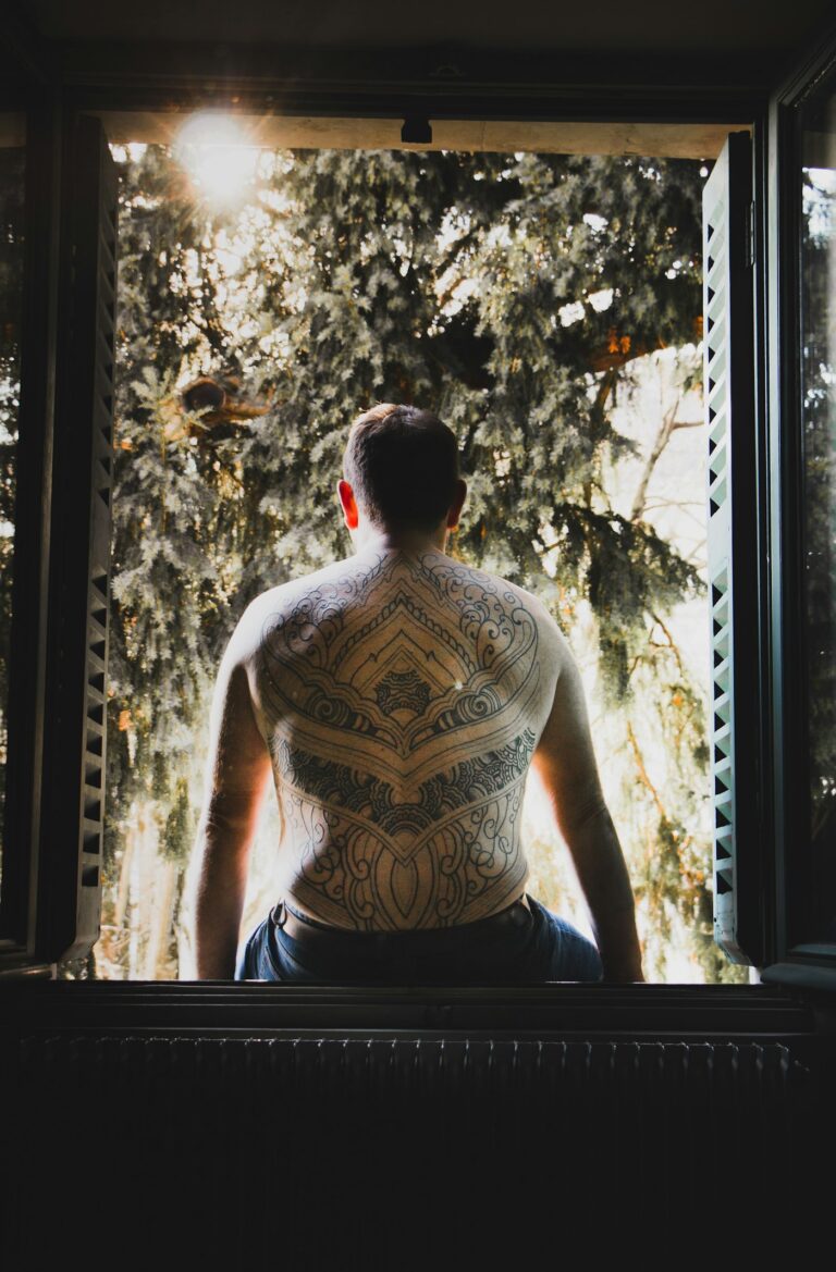Back tattoos for men: man with a full back tattoo sitting in a window