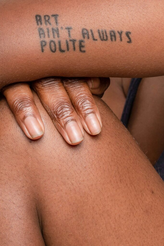 Bold black script tattoo on dark skin reading Art Aint Always Polite