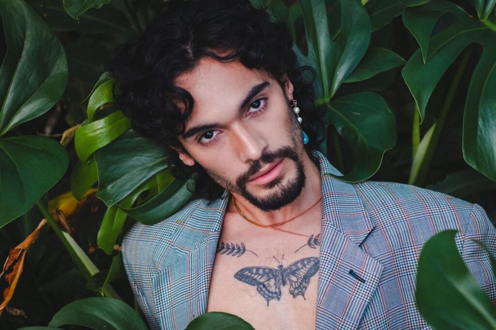 Man in a blazer showing a butterfly tattoo on his chest among monstera leaves